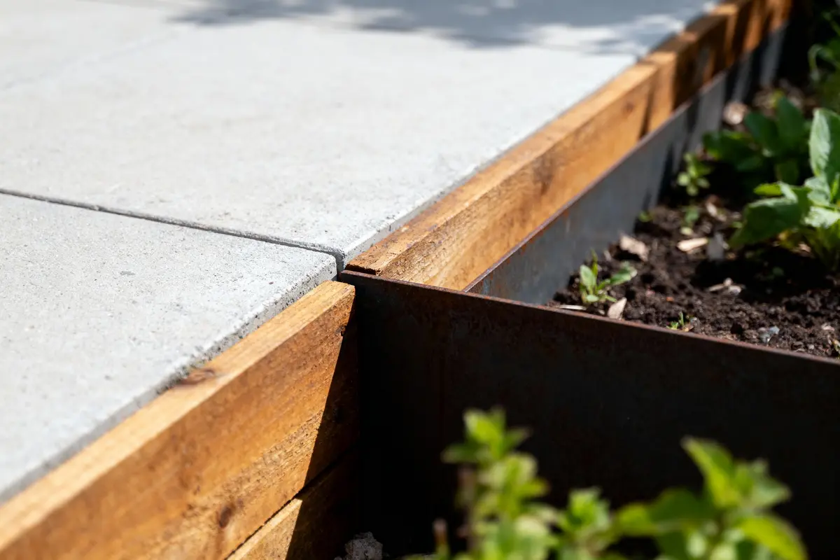 Détail d'un coin de terrasse avec des dalles en béton, bordées de bois et d'un jardin en terre.