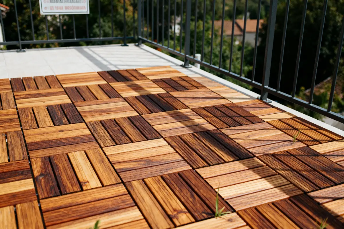 Plancher en bois à motifs alternés, avec des teintes brunes et dorées, sur un balcon. Quelques herbes poussent entre les dalles.