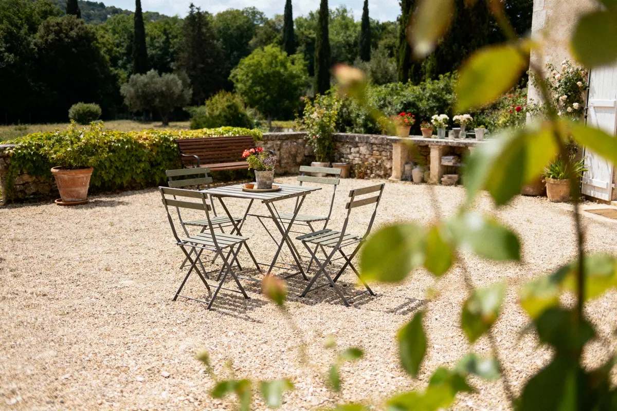 Table et chaises en métal à l'extérieur, entourées de verdure et de fleurs, dans un cadre paisible et ensoleillé.