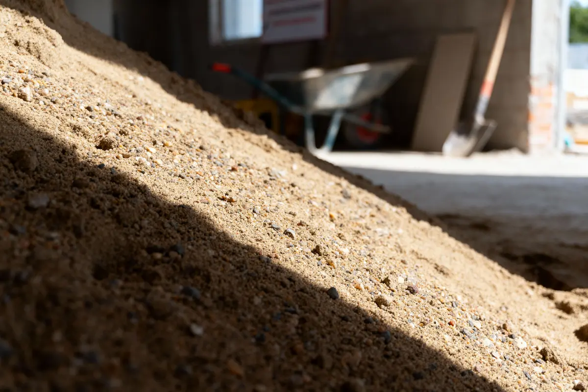 Monticule de sable fin, en premier plan, avec une ombre projetée. En arrière-plan, une brouette et des outils de construction.