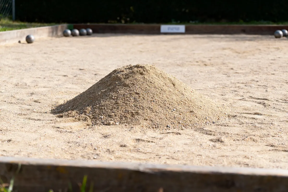 Monticule de sable sur un terrain de pétanque, avec des boules en métal en arrière-plan et un cadre en bois.