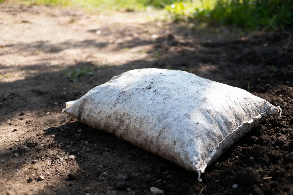 Sac en toile de jute posé sur un sol en terre, légèrement sale, avec des ombres d'arbres en arrière-plan.