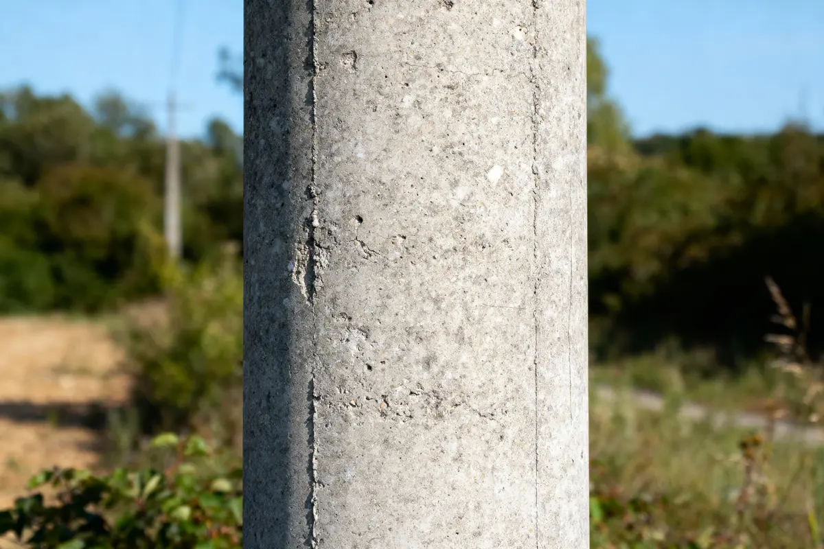 Poteau en béton devant un paysage flou, herbes et arbres à l'arrière-plan sous ciel bleu.