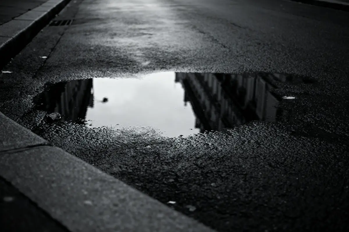 Flaque d'eau sur la chaussée reflétant des bâtiments, ambiance urbaine en noir et blanc.