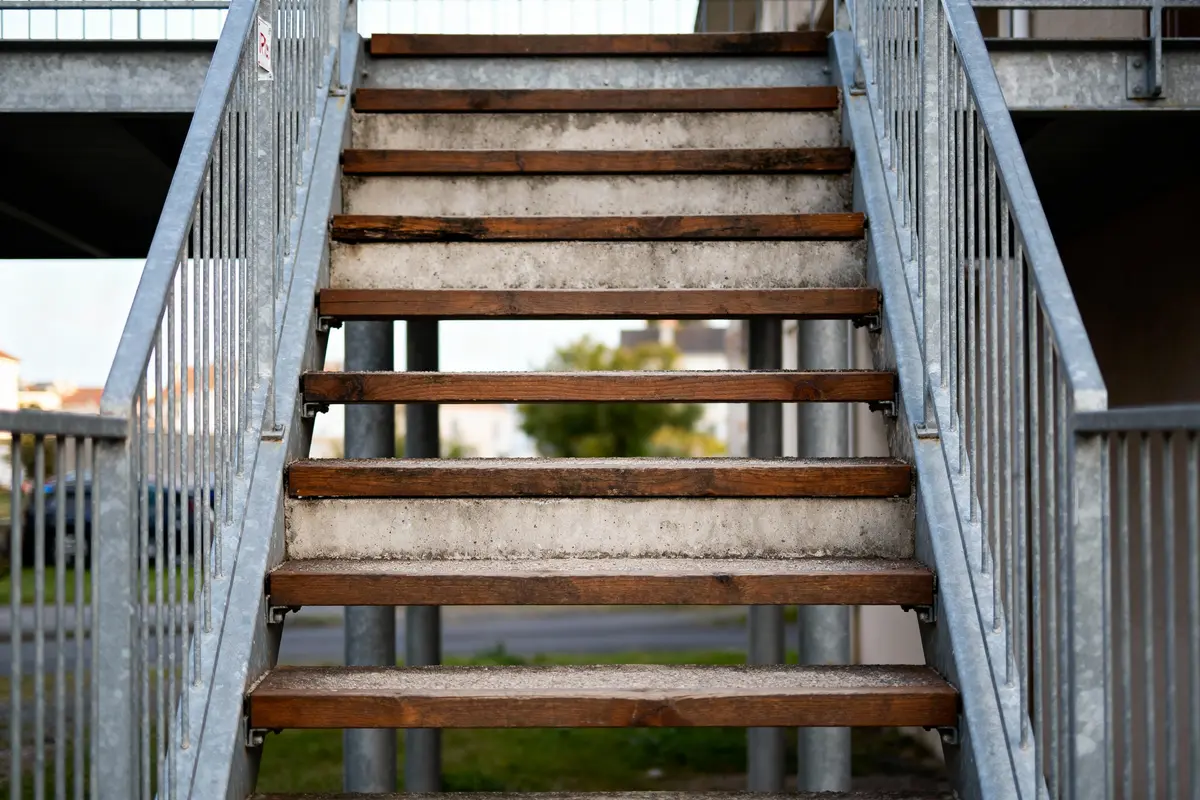 Escalier extérieur, marches en bois, structure métallique, barrière en métal, arrière-plan flou végétation et bâtiment.