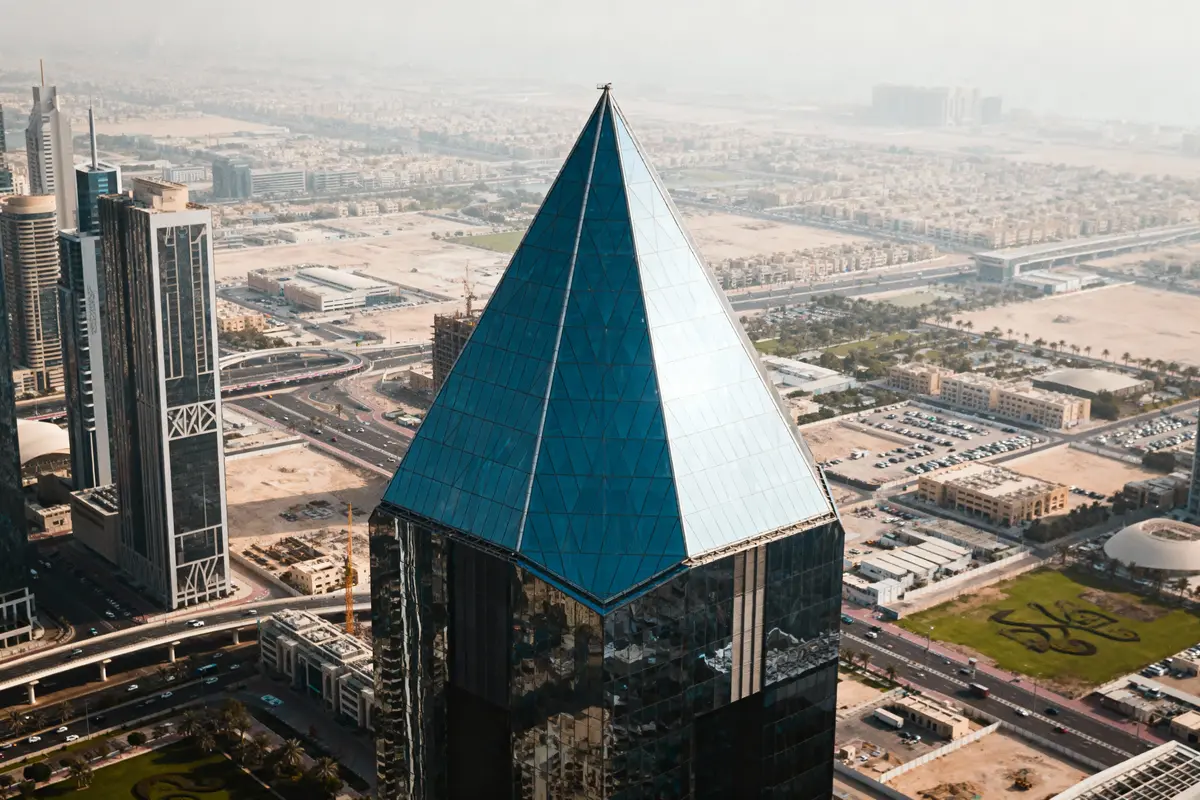 Gratte-ciel moderne à sommet pyramidal en verre, surplombant une ville avec routes et bâtiments environnants.