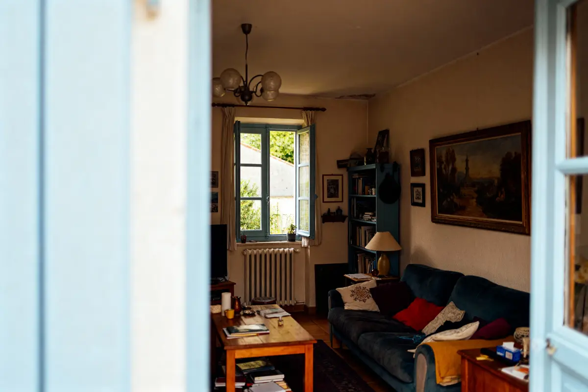 Salon cosy avec canapé bleu, table en bois, fenêtre ouverte, murs décorés, et lumière naturelle.