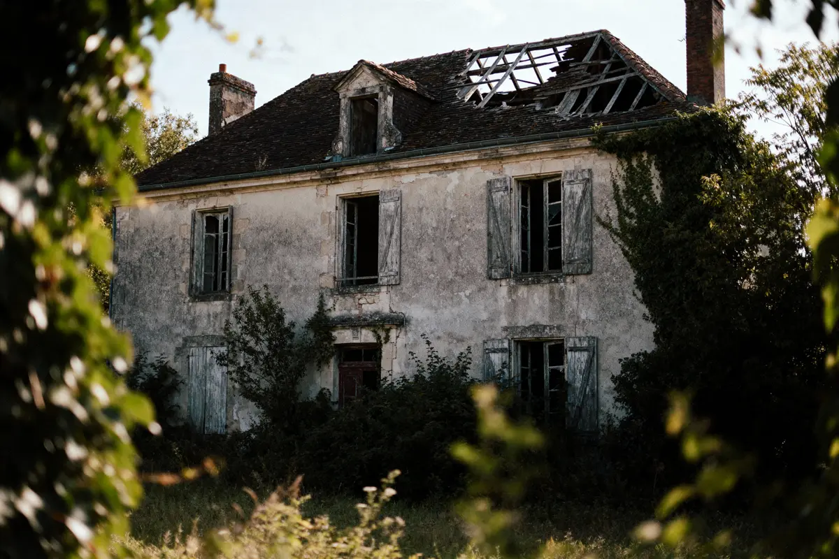 Maison abandonnée, toit endommagé, murs délabrés, fenêtres cassées, envahie par la végétation. Atmosphère sombre et mystérieuse.