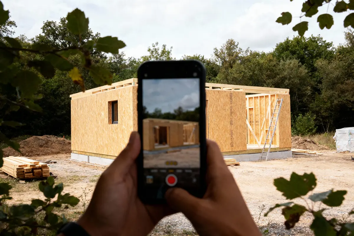 Une personne prenant photo d'une construction en bois, entourée d'arbres, avec téléphone portable.