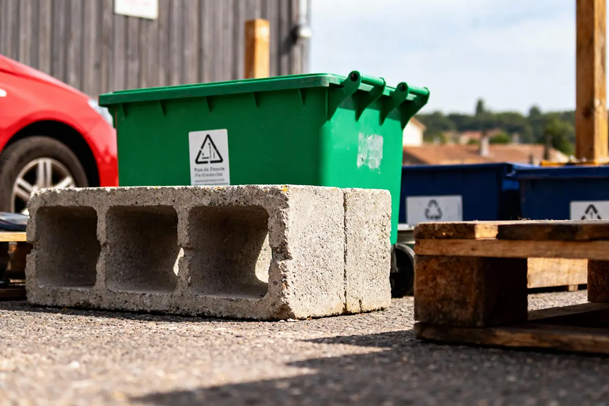Bloc de béton, bac vert et palettes en bois sur sol asphalté près d'une voiture rouge.