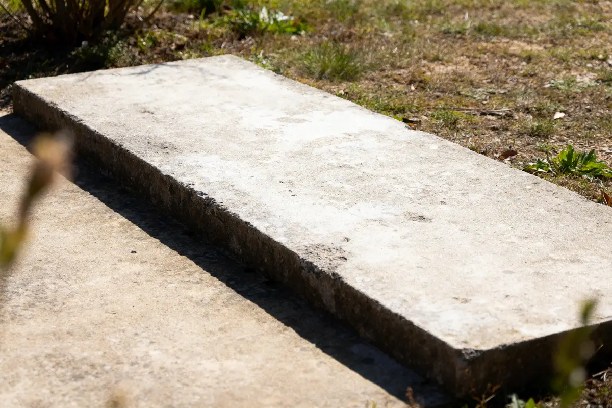 Pierre rectangulaire posée sur le sol extérieur, entourée d'herbe verte clairsemée.