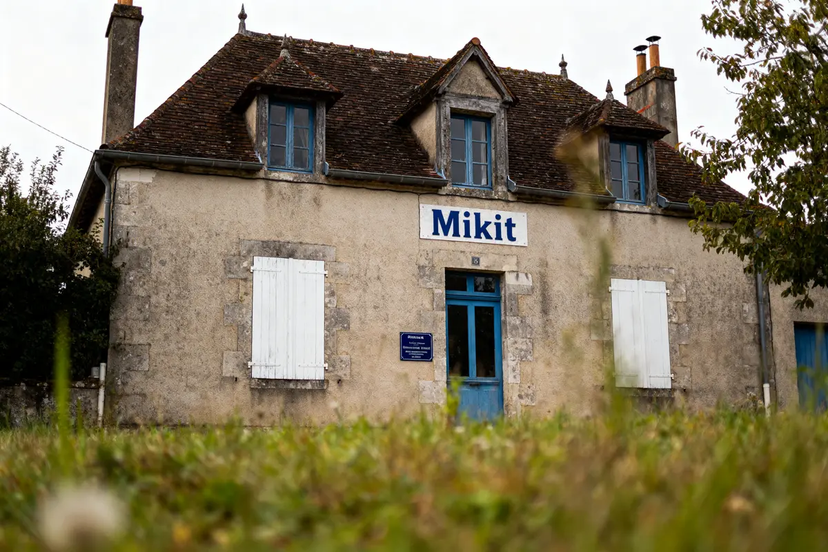 Vieille maison en pierre avec toiture pentue, volets fermés, enseigne “Mikit”, pelouse au premier plan.