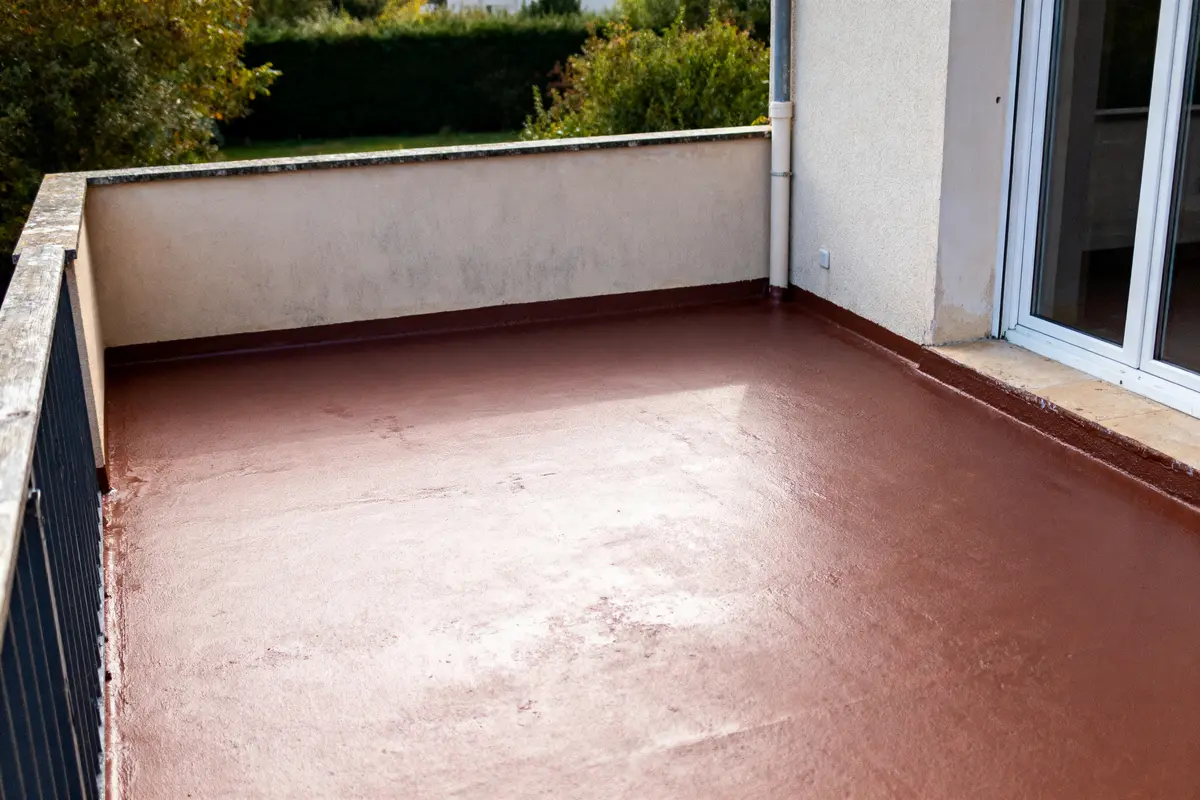 Terrasse en béton brun-rouge avec garde-corps et vue sur végétation environnante, proche d'une porte vitrée.